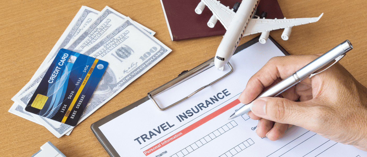 Hand filling out a travel insurance form with credit card, passport, dollars, and airplane model on desk.