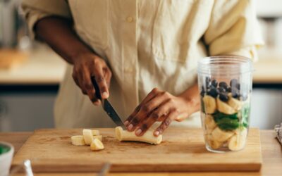 A person slicing a banana on a wooden cutting board, preparing a smoothie with spinach, bananas, and blueberries in a blender cup.