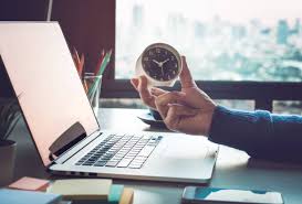 A person holding a small clock in front of a laptop on a work desk, symbolizing time management.