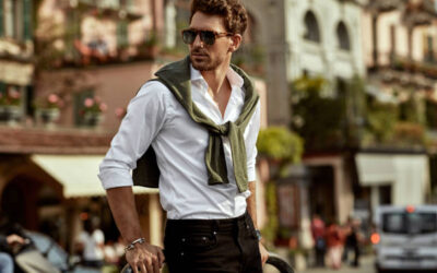Stylish young man in sunglasses wearing a white shirt and sweater over shoulders, showcasing modern street fashion in a European town.