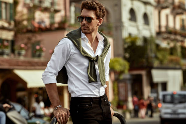 Stylish young man in sunglasses wearing a white shirt and sweater over shoulders, showcasing modern street fashion in a European town.