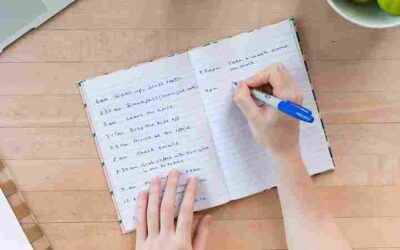 Hand writing a daily schedule in a notebook with a blue pen on a wooden table.