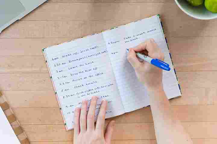 Hand writing a daily schedule in a notebook with a blue pen on a wooden table.