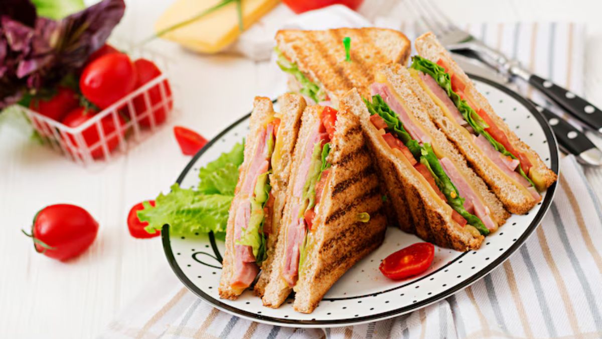 Grilled club sandwiches with lettuce, tomato, ham, and cheese, served on a white plate with cherry tomatoes on the side.