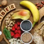 High-fiber diet foods including bananas, pears, raspberries, nuts, seeds, beans, whole grains, and leafy greens arranged on a wooden table.